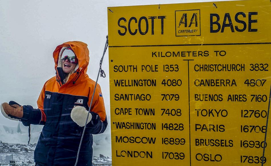 Journalistin Ashlan Gorse Cousteau posiert beim Scott Base Schild in der Antarktis. Das gelbe Schild, im Stil der neuseeländischen Automobile Association, zeigt Entfernungen von Scott Base zu verschiedenen Städten der Welt an. Von hier sind es noch 1353 km bis zum Südpol.